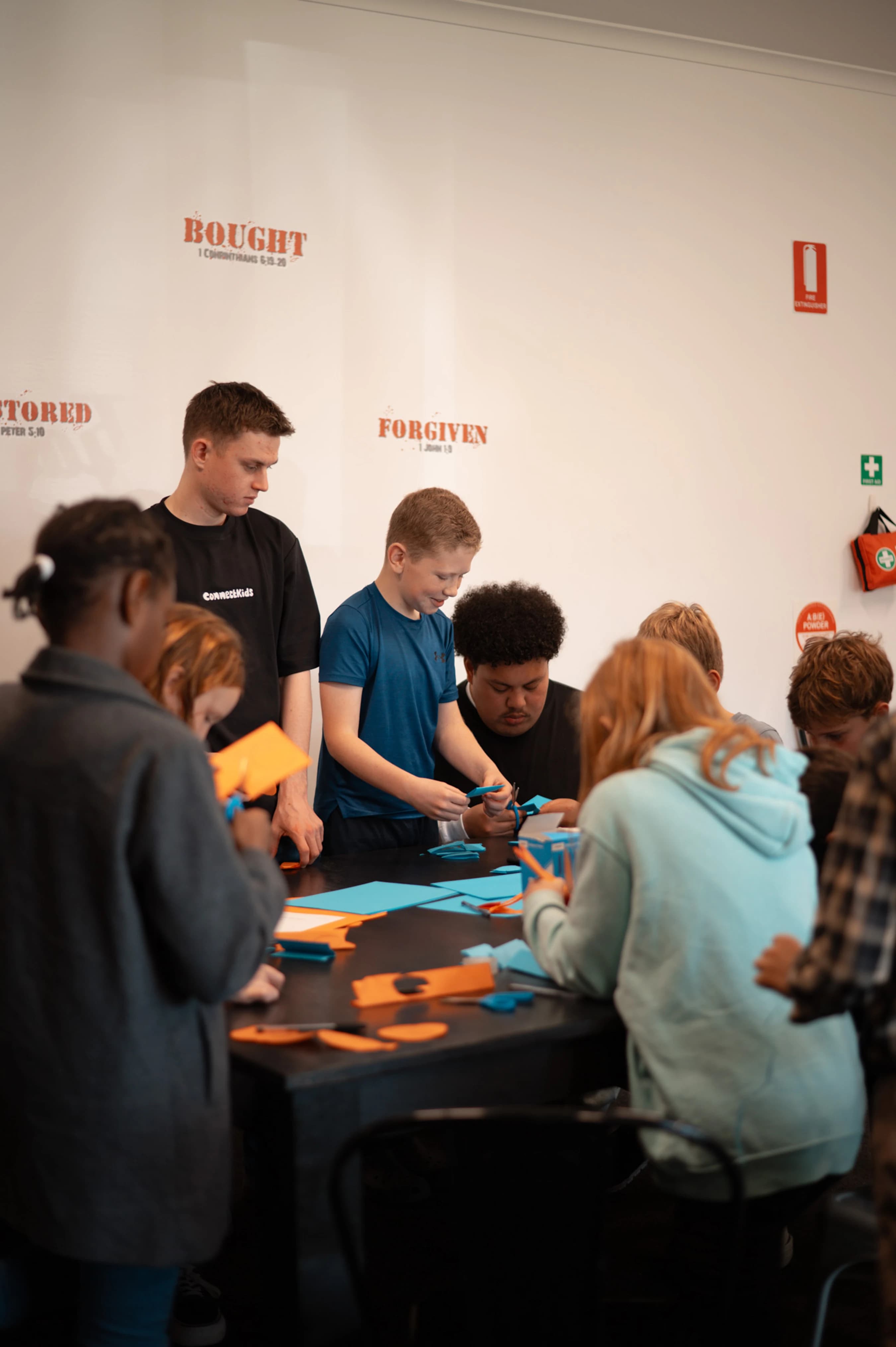A group of diverse children and a young adult leader gather around a table, focused on cutting colorful paper for a craft project in a classroom setting.